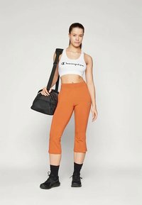 Λευκό κοντό τοπ με το λογότυπο "Champion", πορτοκαλί κολάν 3/4, μαύρα αθλητικά παπούτσια και μαύρη τσάντα γυμναστηρίου. Απλός σχεδιασμός, εφαρμοστή γραμμή.