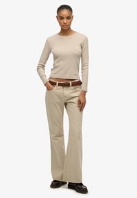 Beige ribstrikket bluse med lange ærmer kombineret med lysebeige vide bukser, fremhævet af et fletbånd i brunt læder. Modellen står selvsikkert iført mørke sko.