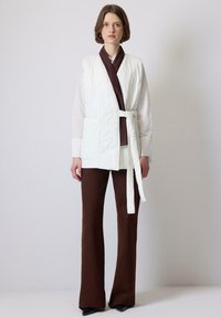 Quilted hvid vest med en brun stribet krave og slips, parret med brune flagrende bukser. Vestens venstre side har en lomme.