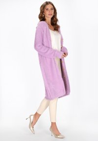Cardigan lungo e soffice di colore lavanda con bottoni dorati, indossato sopra una maglia crema e leggings chiari. Presenta una texture morbida e una vestibilità rilassata.