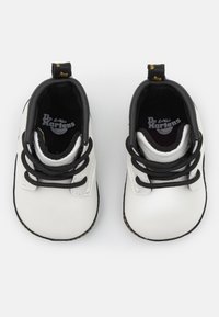 Zapatos para toddlers de cuero blanco con acentos negros, que cuentan con cordones negros, costura amarilla y una pestaña en el talón con el logo visible en su interior.