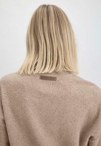 Beiger strukturierter Pullover mit einem gerippten Kragen und einem horizontalen Stoffetikett auf der Rückseite, das ein weiches, gemütliches Aussehen präsentiert.