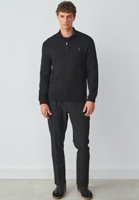 Schwarzer Reißverschluss-Pullover mit einem gerippten Kragen, kombiniert mit schwarzen Hosen und schwarzen Sneakern mit strukturierten Sohlen. Einfaches Logo-Detail auf dem Pullover.