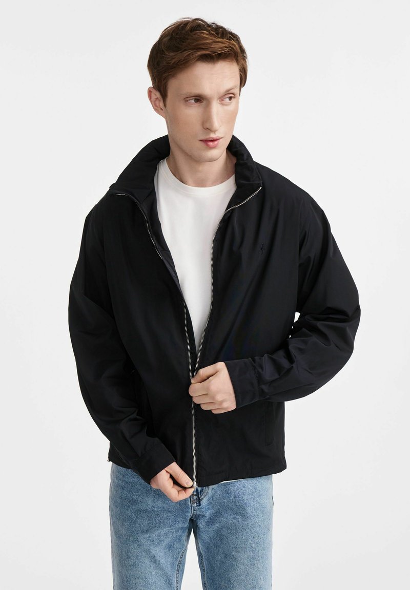 Czarna kurtka na zamek z wysokim kołnierzem, lekki materiał, z długimi rękawami i luźnym krojem. Noszona na białej koszulce.