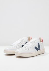 Paire de baskets basses en cuir blanc avec logo "V" bleu marine sur les côtés, lacets blancs, semelles beiges et languettes rouges au talon.