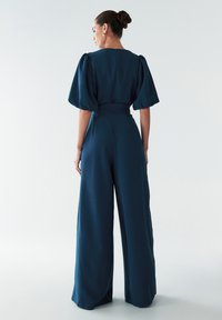 Brede jumpsuit in diep teal, met korte puffmouwen, een aansluitende taille en een verborgen ritssluiting aan de achterkant. Glad, gestructureerd stof.