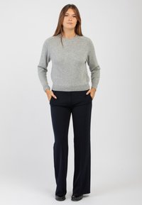 Donna in piedi con le mani nelle tasche, che indossa un maglione grigio, pantaloni neri e scarpe nere, su uno sfondo bianco uniforme.