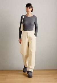 Grå tekstureret langærmet top, cremegul bredbenede bukser med lommer og grå fodtøj; minimalistisk design med subtile detaljer.