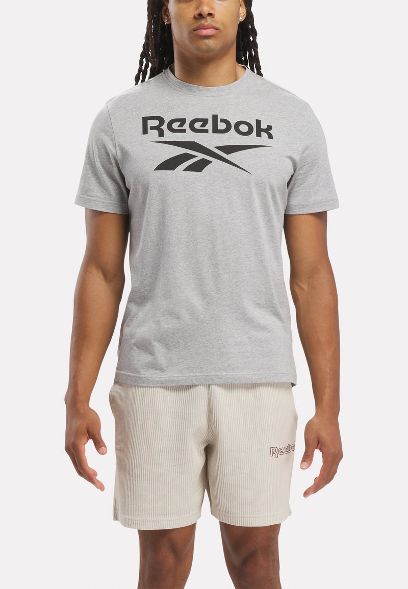 Pelēks īsām piedurknēm T-krekls ar lielu melnu Reebok logo uz krūtīm, apvienots ar gaiši bēšiem ribotiem šortiem ar mazu sarkanu logo.