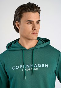 Felpa verde con coulisse, caratterizzata da un logo frontale in bianco e azzurro chiaro. Tessuto morbido, vestibilità rilassata e tasca a marsupio.