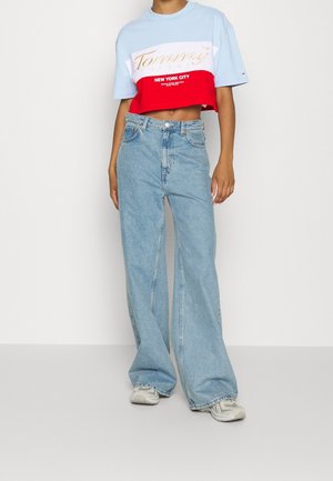 Person iført en farveblokeret, cropped Tommy Hilfiger t-shirt og vidde bukser i lyseblå med hvide sneakers mod en ensfarvet baggrund.