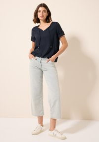 Top navy a maniche corte con scollatura a V e dettagli con cordino, abbinato a jeans a gamba larga di colore azzurro chiaro e sneakers bianche con accenti verdi.