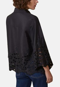 Blusa nera con collo a camicia, maniche a campana e motivi floreali intagliati sull'orlo. Texture liscia e vestibilità ampia.