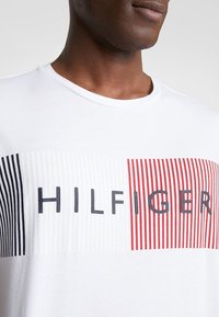 Bílý bavlněný tričko s vodorovným logem "HILFIGER" s červenými a námořnickými pruhy. Klasický kulatý výstřih a krátké rukávy.