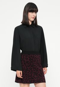 Mujer con cabello oscuro y lacio, vistiendo una camisa negra de manga larga y una falda de estampado de leopardo púrpura, de pie contra un fondo blanco.
