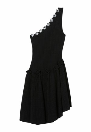Grande femme portant une robe noire asymétrique avec des détails floraux, tenant un sac à main noir et portant des chaussures slip-on noires avec des semelles imprimées.