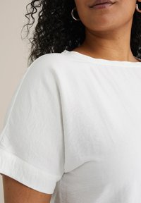 Top blanco de manga corta con un ajuste relajado y una textura ligeramente visible. Presenta un escote ancho y puños enrollados para un aspecto casual.
