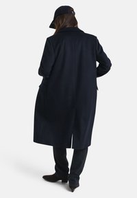 Person wearing a long dark coat, black pants, and dark ankle boots, standing with back facing camera, also wearing a dark baseball cap.