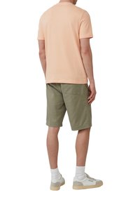 Licht perzikkleurig katoen t-shirt met korte mouwen, gecombineerd met olijfgroene shorts. Model draagt witte sneakers met crèmekleurige accenten en geribbelde sokken.