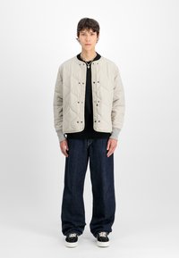 Jauns cilvēks, tērpies gaiši bežā piepildītā jakā, melnā krekliņā, tumšās plašajās bikses, melnās un baltās sporta kedās, stāv uz neitrālas baltas fona.