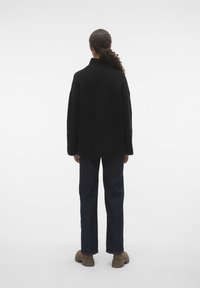 Svart oversized genser med høy krage, strikket tekstur og senkede skuldre, kombinert med mørk denim jeans og brune sko.