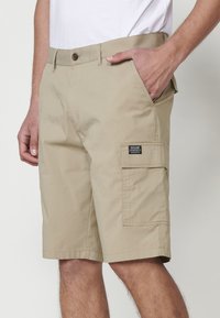 Shorts cargo bej de țesătură ușoară, cu un buzunar lateral prevăzut cu o etichetă și o închidere clasică cu nasture la talie.