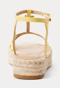 Žlutá sandála na kotníkovém pásku s tkanou juty na platformě, béžová stélka a zlatá přezka, zobrazená zezadu na bílém pozadí.