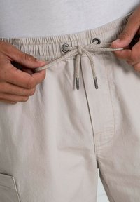 Pantalones cortos beige claro con cintura elástica y cordón. La tela tiene una textura sutil y cuenta con un detalle de bolsillo lateral.