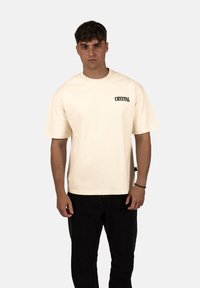 Krémové bavlněné tričko s krátkými rukávy, kulatým výstřihem a potiskem "CRYSTAL" na levém prsu. Kombinováno s černými kalhotami.