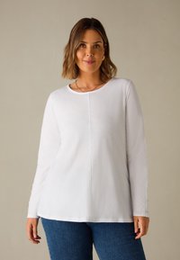 Maglietta bianca a maniche lunghe con scollatura rotonda, bottoni laterali e vestibilità rilassata, abbinata a jeans in denim blu. Morbido tessuto di cotone.