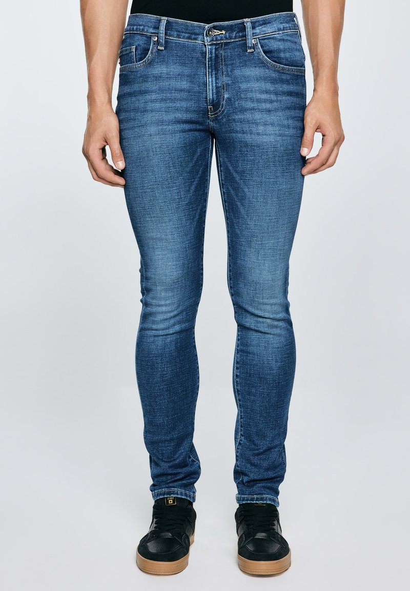 Egy férfi, aki szűk szabású kék farmert és fekete gumitalpú tornacipőt visel, egyszínű világos háttér előtt áll.