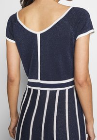 Mörkblå stickad topp med vita detaljer, med en V-ryggdesign, korta ärmar och ett randigt mönster på den nedre delen. Mjuk textur med lite lyster.