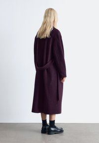 Langer, dunkelvioletter Mantel mit strukturierter Oberfläche, ausgestattet mit einem Gürtel, Taschen an den Seiten und langen Ärmeln. Getragen mit schwarzen Schuhen und Socken.