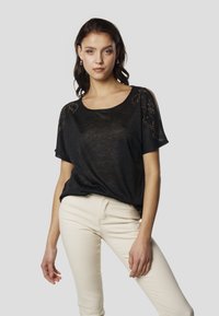 Fekete rövid ujjú póló, széles nyakkivágással és díszítő gyöngyberakással a vállakon. Világos színű, testhez simuló nadrággal viselve.