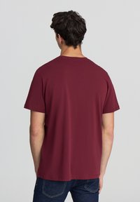 Maglietta a maniche corte bordeaux con scollo rotondo e texture liscia, abbinata a jeans blu scuro. La vista posteriore mostra un design semplice e una vestibilità comoda.