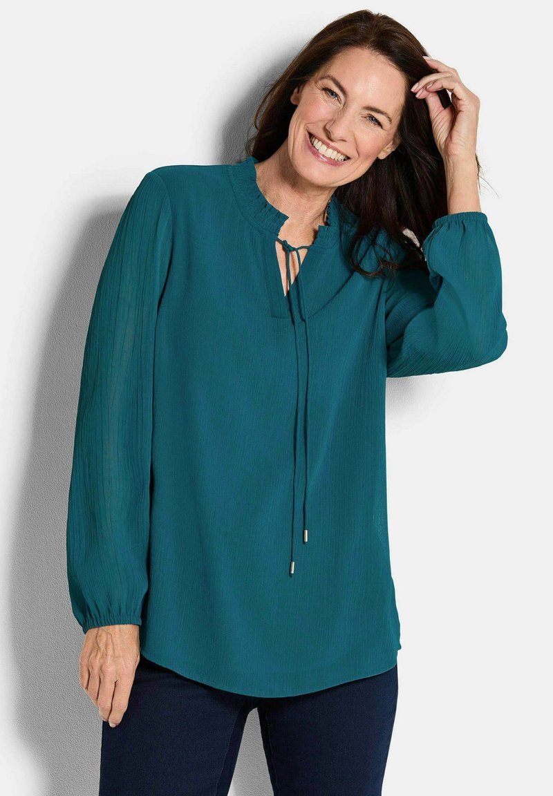 Glunderende vrouw die haar aanraakt, gekleed in een turquoise blouse met lange mouwen en strik, met donkere broeken, staande tegen een lichte achtergrond.