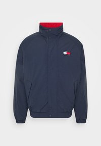 Marineblauw windjack met hoge kraag, rood binnenvoering, drukknoopsluitingen aan de voorkant, zijzakken en een klein logo op de borst.