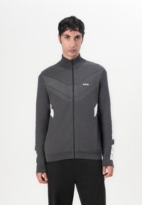 Dunkelgrau zip-up Jacke mit hohem Kragen, die mit strukturierten Mustern und weißen seitlichen Akzenten ausgestattet ist. Hergestellt aus weichem, dehnbarem Stoff.