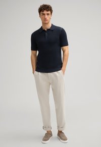 Marineblauwe polo met korte mouwen en een textuurpatroon, gecombineerd met lichtbeige broeken en tan instap schoenen met een patroon.