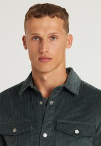 Donkergroene corduroy overhemd met een button-down kraag, zilveren drukknoopknopen en twee borstzakken. Verticale ribbeltextuur.
