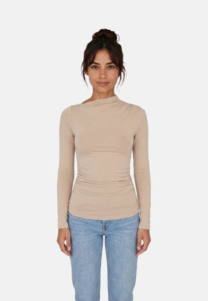 Donna con capelli scuri raccolti in uno chignon, indossa una maglia beige a maniche lunghe e jeans azzurri, in piedi davanti a uno sfondo bianco semplice.