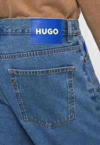 Τζιν παντελόνια σε ανοιχτό μπλε χρώμα, με ένα μεγάλο μπλε λάβαρο με την επιγραφή "HUGO" στη μέση και μια πίσω τσέπη με αντίθετη ραφή.