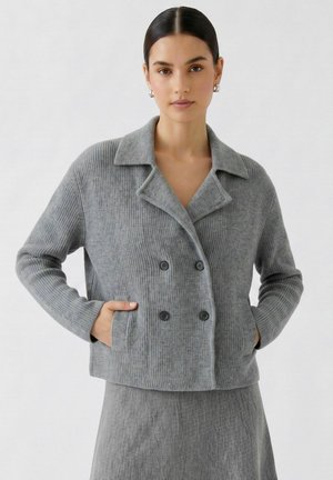 Frau mit dunklen Haaren in einem niedrigen Dutt, trägt silberne Creolen und eine graue, gerippte, doppelreihige Jacke mit passendem Rock.