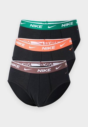 Homme mince et musclé portant un slip noir Nike, debout pieds nus devant un fond clair uni, les mains derrière le dos.