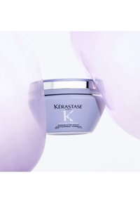 Kérastase Blond Absolu violette Haarmaske im Glas mit silbernem Deckel, umgeben von weichen Lavendelballons vor weißem Hintergrund.