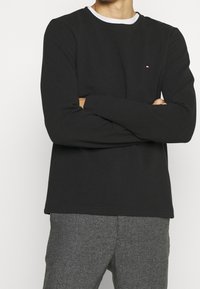 Schwarzes Langarmshirt mit weißem Rundhalskragen, gerippter Textur und einem kleinen Logoakzent auf der Brust. Kombiniert mit grau karierter Hose.