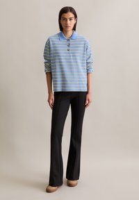 Polo rayé en bleu clair avec des rayures vertes et blanches, col boutonné et coupe ample, associé à un pantalon évasé noir et des chaussures beiges.