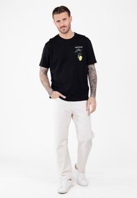 Man met getatoeëerde armen, gekleed in een zwart T-shirt met een citroendesign, witte broek en witte sneakers, staande voor een witte achtergrond.