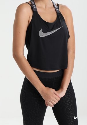 Frau trägt schwarzes Nike athletisches Croptop und hoch taillierte Leggings, steht mit vor der Brust verschränkten Händen vor einem schlichten Hintergrund.