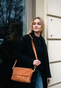 Woman in black coat carrying brown leather shoulder bag, standing by beige wall with glass reflection behind her.